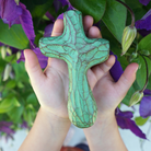 Hands holding patina green painted wooden holding cross in front of purple flowering plant